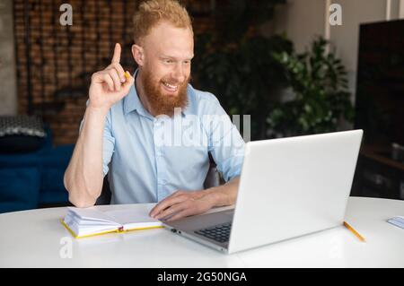 Un giovane uomo d'affari entusiasta con capelli rossi in una camicia casual intelligente che condivide idee durante la conversazione video sul laptop, un uomo che tiene il dito indice in alto e guarda lo schermo del laptop, discutendo online Foto Stock