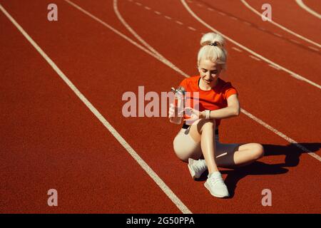 giovane bella bionda si siede su una pista da jogging con una bottiglia d'acqua e guarda il suo orologio Foto Stock