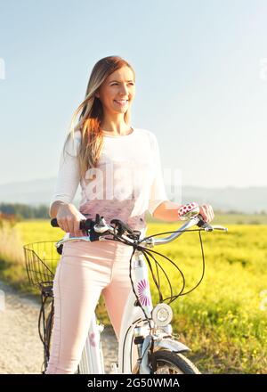 Giovane donna in piedi accanto alla sua bici su strada di campagna polverosa, forte luce del sole pomeriggio in background splende su campo di fiori gialli Foto Stock