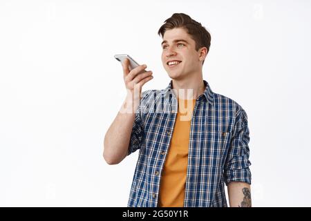 Immagine di un giovane sorridente che parla con il vivavoce, registra messaggi vocali o traduce spach con l'app per telefono cellulare, in piedi casual contro il bianco Foto Stock