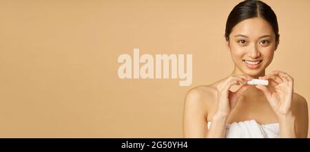 Intestazione sito web di Ritratto di adorabile giovane donna avvolto in asciugamano sorridente mentre tenendo balsamo labbro, in posa isolato su sfondo beige Foto Stock