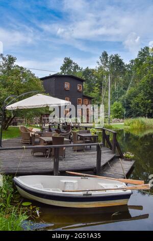Tavoli da ristorante situati su un molo, vecchio, rinnovato mulino ad acqua e una vecchia, piccola barca in piedi vicino al lago, foresta sullo sfondo Foto Stock