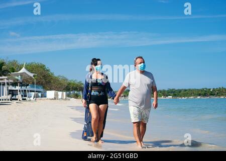 Coppia matura con maschera protettiva che tiene le mani e passeggiando sulla spiaggia Foto Stock