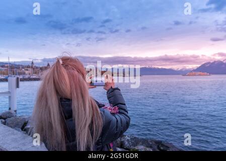 Donna bionda non riconosciuta che usa il suo smartphone per scattare una foto della costa durante il tramonto. Foto Stock