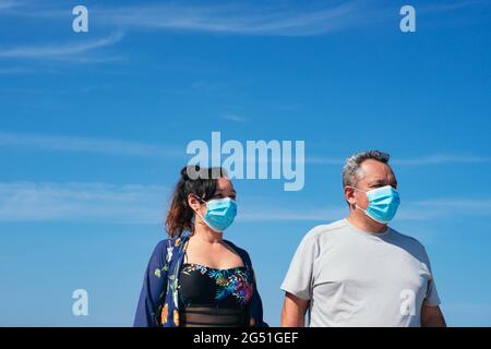 Coppia matura con maschera protettiva che tiene le mani e passeggiando sulla spiaggia Foto Stock