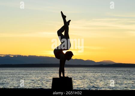 Silhouette di donna che fa il supporto acrobatico posa contro il mare al tramonto Foto Stock