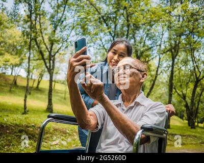 Nonno in sedia a rotelle e nipote che parlano videochiamata con i parenti tramite smartphone nel parco. Vita familiare in vacanza. Foto Stock