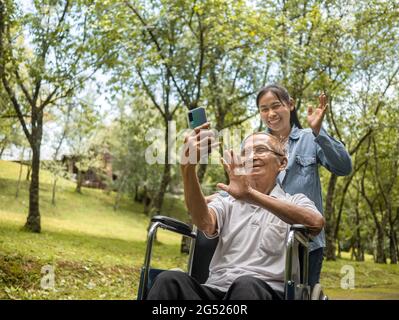 Nonno in sedia a rotelle e nipote che parlano videochiamata con i parenti tramite smartphone nel parco. Vita familiare in vacanza. Foto Stock