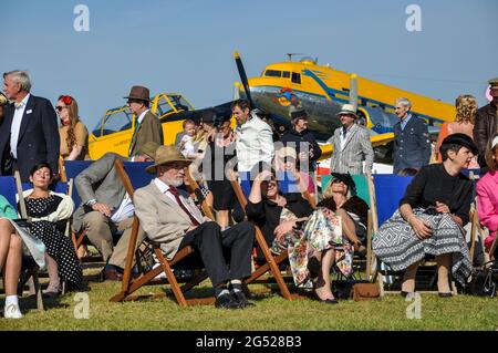 I membri del pubblico al Goodwood Revival 2012 vestiti in abito d'epoca, seduti intorno su sedie a sdraio in tempo di sole. Piani dietro Foto Stock