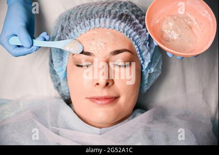 Primo piano della mano del cosmetologo in guanto sterile blu che applica la maschera cosmetica sul viso femminile del cliente. Giovane di charme che ha la procedura di cura della pelle in salone di bellezza. Concetto di cura della pelle, bellezza e benessere. Foto Stock