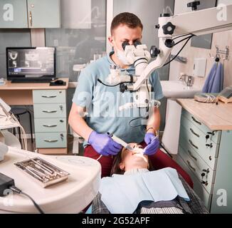 Giovane dentista maschile che usa strumenti per microscopio e dentisti mentre si trattano i denti della donna. Paziente femminile sdraiato in sedia dentale durante il trattamento dentale in centro medico. Concetto di odontoiatria. Foto Stock