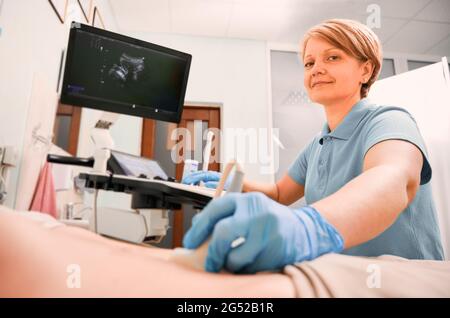 Donna ecografa che esamina il paziente con uno scanner a ultrasuoni. Medico che guarda la fotocamera e sorride durante l'esame ecografico. Concetto di assistenza sanitaria, ecografia e diagnostica a ultrasuoni. Foto Stock