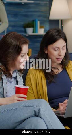 Donne multietniche che si divertono insieme a guardare un film sul computer portatile mentre si riposano sul divano. In background donna e uomo bevono birra, ridendo a tarda notte durante la festa rilassante Foto Stock