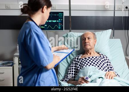 Assistente medico che controlla il trattamento dell'uomo anziano che legge le note sulla clipboard. Ammalato anziano che si trova a letto discutendo con l'infermiere circa l'esperienza di diagnosi con la sacca per infusione attaccata. Foto Stock