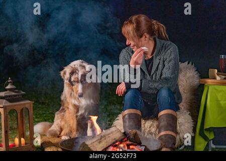 Giovane donna sta mangiando una fetta di baguette con brie. Il suo giovane cane Pastore australiano si siede accanto a lei, guardando il falò con il suo cucciolo blu Foto Stock
