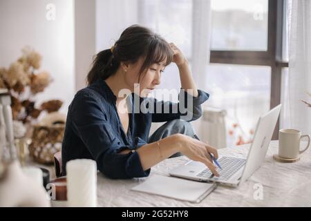 Giovane donna asiatica pensiva freelance in abbigliamento casual che lavora su laptop a distanza da casa o studiare online, donna che guarda lo schermo e prendere appunti Foto Stock