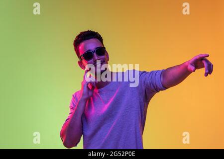 Ritratto del giovane latino in occhiali da sole che si posano isolati su sfondo giallo verde sfumato in luce al neon. Foto Stock