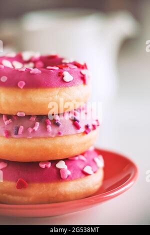 Su un piatto rosso luminoso sono tre deliziose ciambelle rosa con spruzzi sotto forma di cuori, e c'è una teiera bianca sullo sfondo. una rom Foto Stock