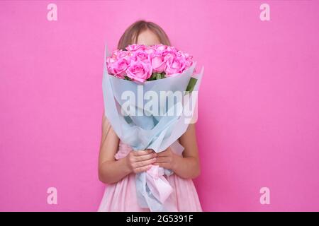 Una bambina in abito rosa con bouquet di rose, nascose dietro il flowers.The ragazza inala la fragranza di fiori su sfondo rosa Foto Stock