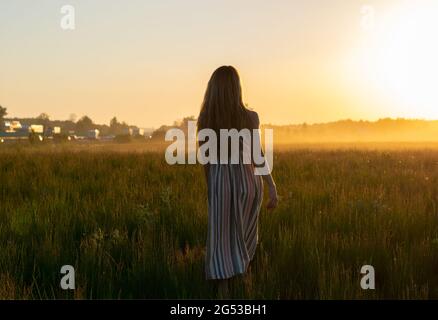 una ragazza in un vestito cammina attraverso un campo con nebbia al tramonto verso il sole Foto Stock