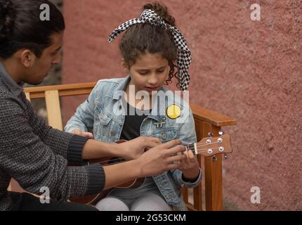 Giovane uomo ispanico insegnamento ragazza capelli ricci per giocare ukulele Outdoors musica concetto Foto Stock