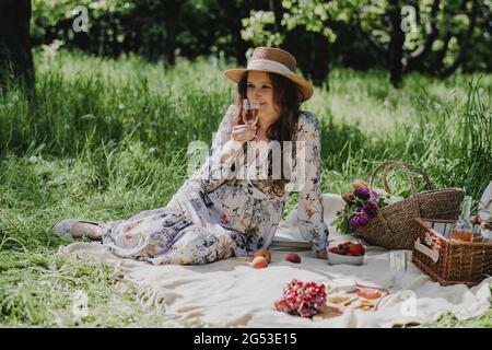 Giovane donna in abito estivo e cappello di paglia seduta con vino rosato, formaggio, frutta fresca e baguette su coperta. Foto Stock