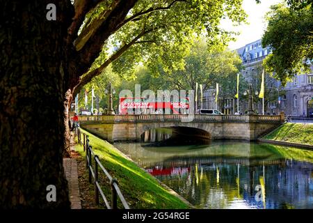 The beautiful city canal "Stadtgraben" on Königsallee with its old trees. Red doubledecker tour bus waiting on a bridge crossing the canal. Foto Stock