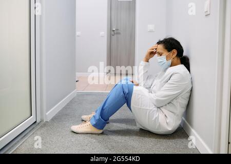 Dopo una dura giornata di lavoro in una clinica medica, stanca una dottoressa si siede sul pavimento in un ospedale di corridoio con gli occhi chiusi, indossando una maschera medica. Pand Foto Stock