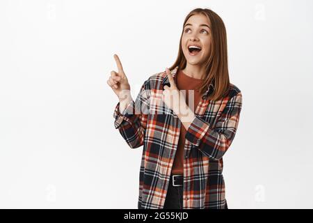 Pubblicità e concetto di gente. Happy girl che punta, guardando l'angolo in alto a sinistra con sorriso eccitato, controllare i prezzi del negozio, mostra il logo o la promozione Foto Stock