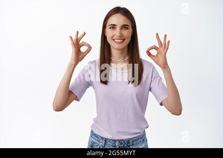 Immagine di sorridente giovane donna mostra OK OK segni, zero problemi, raccomandare qualcosa di buono, feedback positivo, mi piace e concorda, approva, in piedi Foto Stock