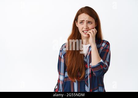 Studentessa insicura con i capelli rossi, si sente ansioso, guardando a sinistra banner con faccia spaventato, mordendo le dita e diventando nervoso, in piedi contro bianco Foto Stock