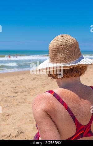 Una donna adulta in costume da bagno e un cappello si siede in riva al mare in una giornata di sole, vista posteriore. Vacanze estive, viaggi e turismo. Foto Stock