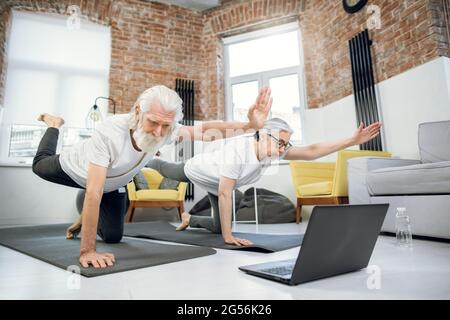 L'uomo anziano e la donna si equilibrano sul tappetino yoga su una mano e sul ginocchio mentre si allungano gli altri sui lati opposti. Coppia senior che utilizza un computer portatile moderno per la formazione a casa. Foto Stock