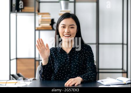 Webcam headshot di una giovane donna asiatica, freelance o manager, positiva e sicura, si siede alla scrivania, parla con colleghi o amici tramite collegamento video, agitando la mano, sorridendo amichevole Foto Stock