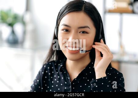 Primo piano di una giovane donna asiatica con capelli lunghi, piacevole e sicura in cuffia, freelance, operatrice o consulente di call center, indossando una camicia nera elegante, guarda direttamente alla fotocamera, sorride amichevole Foto Stock