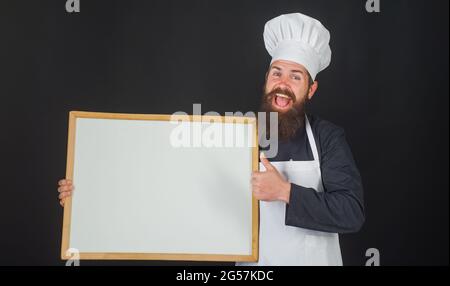 Chef con menu a tavola vuoto. Il cuoco sorridente o il panettiere mostra il pollice in su. Pubblicità. Spazio di copia per il testo. Foto Stock