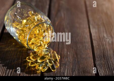 Vitamine per la salute e la longevità sotto forma di capsule di gelatina gialla sparse da un vaso di vetro su un tavolo di legno. Foto Stock