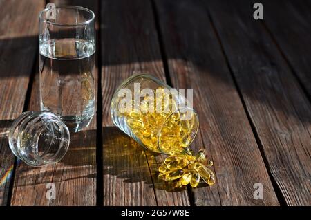 Supplemento dietetico in capsule di gelatina gialla in un vaso di vetro accanto ad un bicchiere d'acqua su un tavolo di legno. Vitamine per la salute e la longevità. Foto Stock