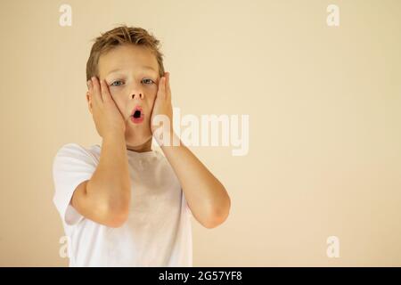 Un ragazzo sorpreso o spaventato di 7-10 anni in una T-shirt bianca si alza e grida con le mani sulle guance su uno sfondo beige. Spazio di copia. Foto Stock