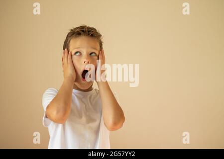 Un ragazzo sorpreso o spaventato di 7-10 anni in una T-shirt bianca si alza e grida con le mani sulle guance su uno sfondo beige. Spazio di copia. Foto Stock