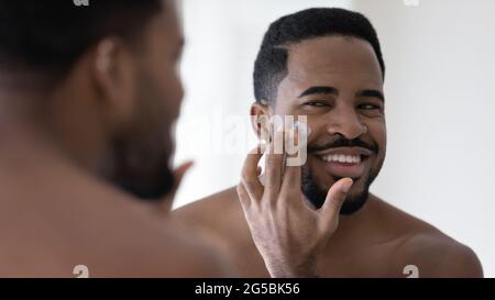 Felice bell'uomo afro-americano che applica idratante sul viso Foto Stock