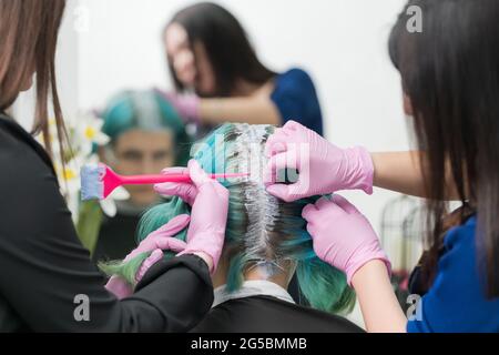 Processo di tintura dei capelli in salone di bellezza professionale. Due acconciatori femminili applicano la vernice ai capelli della donna durante la decolorazione delle radici dei capelli. Foto Stock