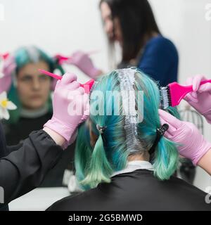 Processo di tintura dei capelli in salone di bellezza. Due parrucchieri professionali in guanti protettivi medici rosa che applicano vernice ai capelli femminili durante la decolorazione Foto Stock