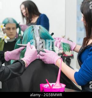 Processo di tintura dei capelli in salone di bellezza. Due parrucchieri femminili in guanti protettivi rosa che applicano vernice ai capelli durante la decolorazione delle radici dei capelli. Foto Stock