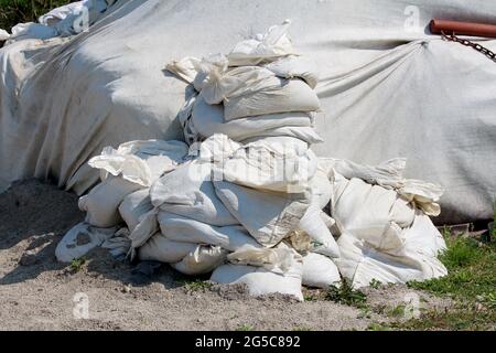 Pile di nuovi sacchi di sabbia bianchi utilizzati per la protezione contro le alluvioni accanto alla parete temporanea di protezione contro le alluvioni realizzata con barriere scatolanti rivestite con tessuto geotessile spesso Foto Stock