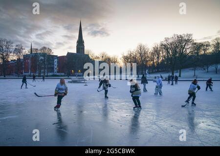 I giocatori di hockey su ghiaccio e alcuni pattinatori di figura pattinano sul laghetto congelato durante l'inverno a Queens Park, Glasgow, Scozia, Regno Unito Foto Stock