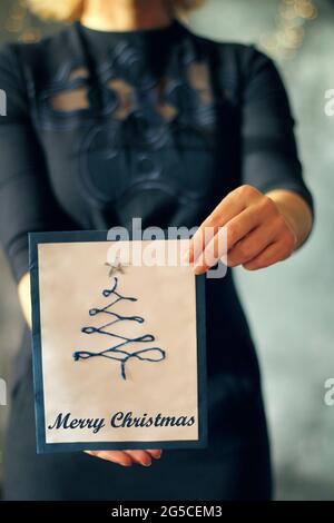 Ragazza che tiene una carta dell'albero di natale davanti a lei, faccia coperta, fuoco selezionato sulla carta e le mani, concetto di Natale Foto Stock