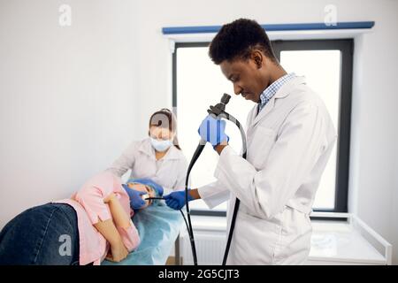 Esame endoscopico in clinica medica moderna. Giovane sicuro medico afroamericano maschio tiene un endoscopio in mano, inserendo la fotocamera nella bocca della paziente femminile Foto Stock