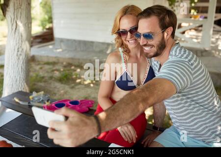 giovane coppia adulta caucasica che prende selfie, seduto su una panchina nel cortile, sorridente Foto Stock