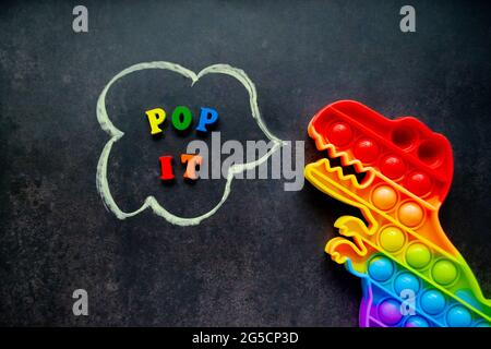 Pop it dinosauro giocattolo arcobaleno colori su uno sfondo nero con lettere multicolore e l'iscrizione - Pop it in una bolla di parola. Foto Stock